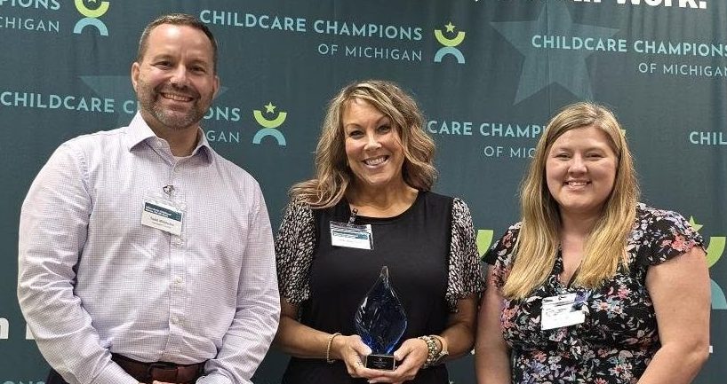 Three people standing in front of a backdrop that reads 'Childcare Champions of Michigan'. The woman in the center holds an award trophy.