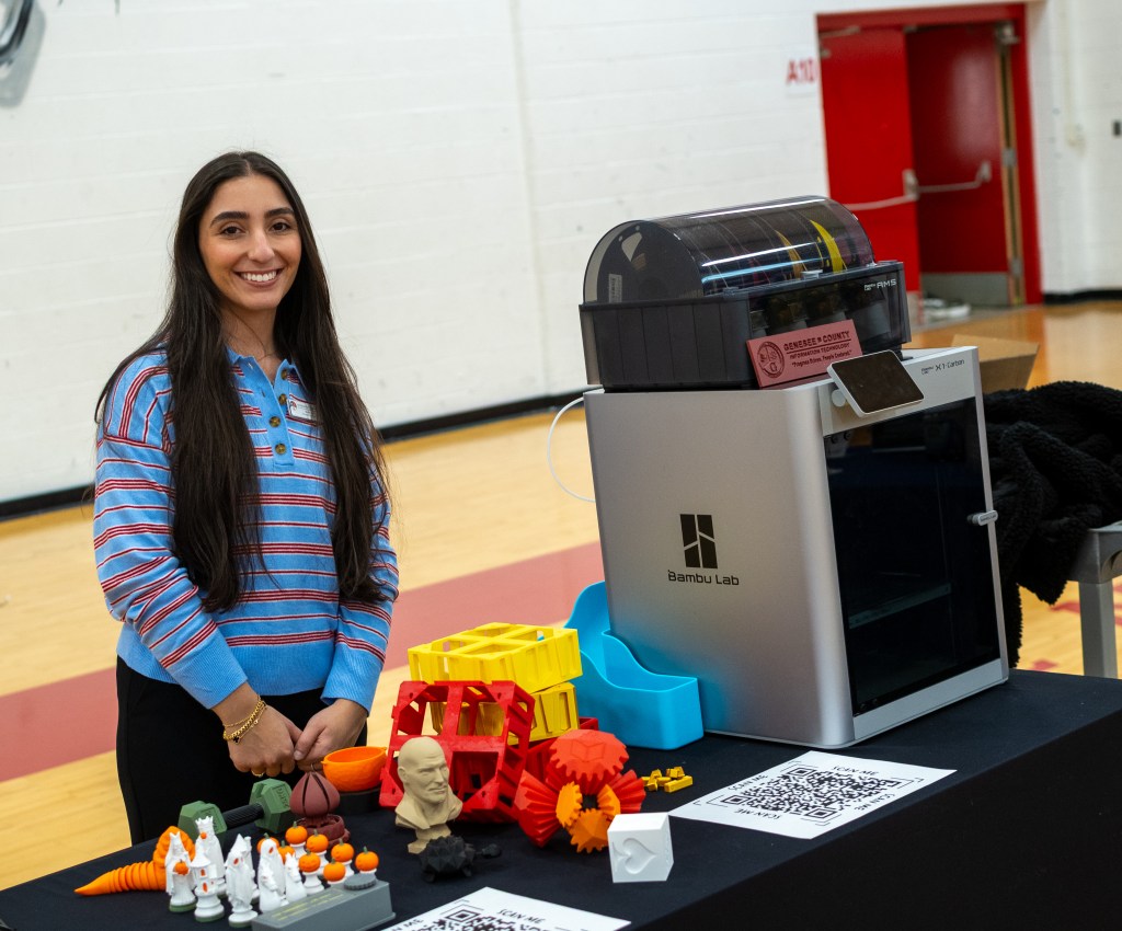 A woman with long hair smiles beside a Bambu Lab 3D printer, showcasing various 3D printed objects on a table, including colorful toys and figurines.