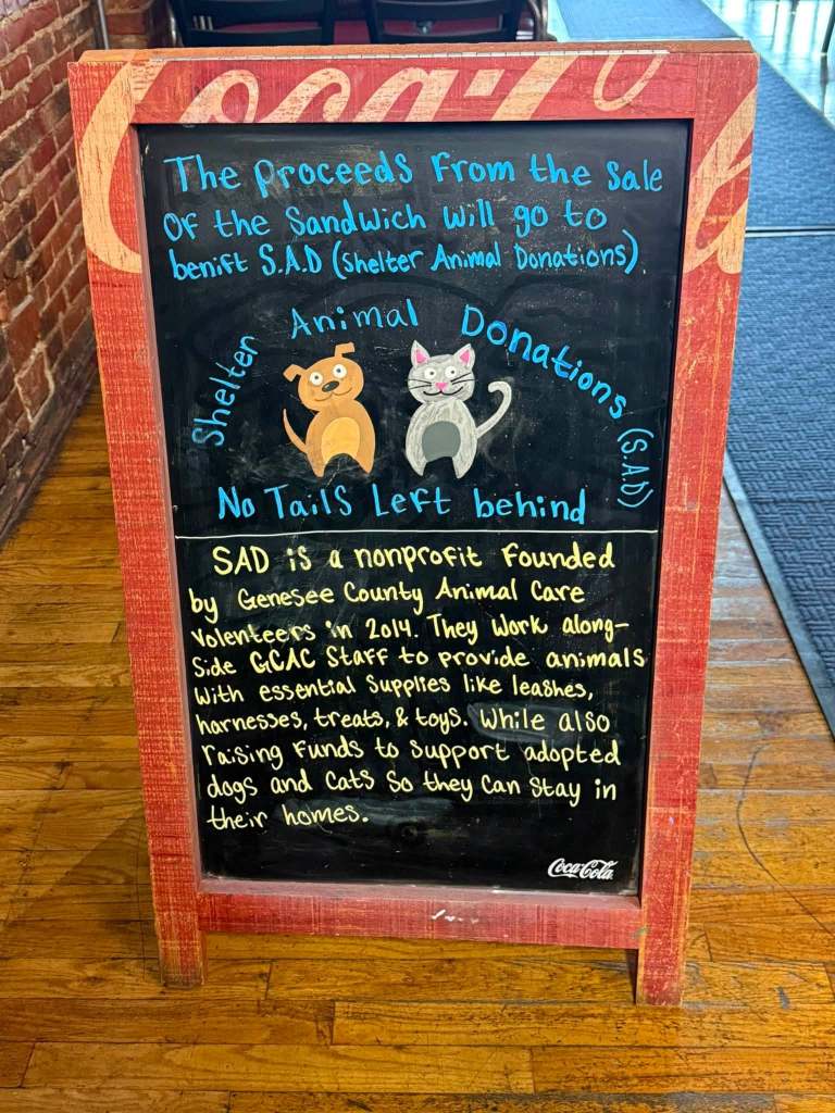 Chalkboard sign promoting the sale of a sandwich, stating proceeds will go to Shelter Animal Donations (SAD). The sign describes SAD as a nonprofit that supports animals in need.