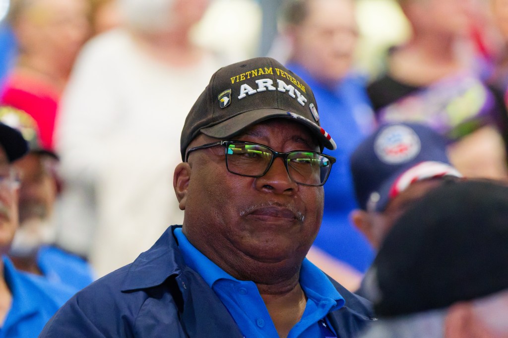 A middle-aged African American man wearing a navy blue shirt and a black cap that reads 'Vietnam Veteran Army' with glasses, looking serious in a crowd of people.