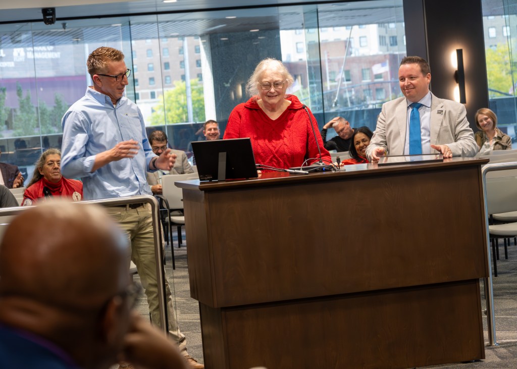 A group of three people standing at a podium, with two men on either side of a woman in a red sweater, smiling during a presentation. In the background, an audience is visible, engaged and attentive.