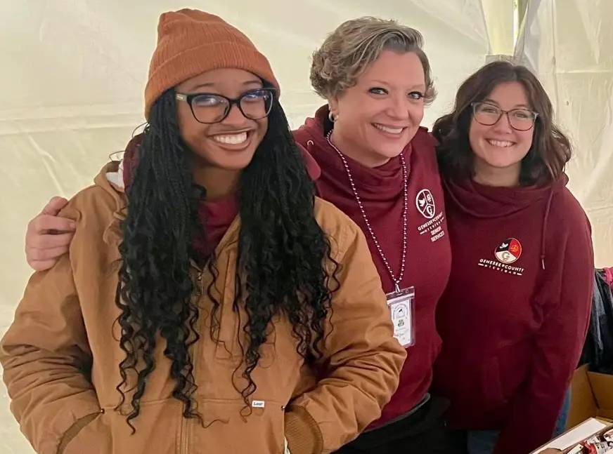 Three women smiling together, wearing casual clothing and cozy sweaters, standing under a tent.