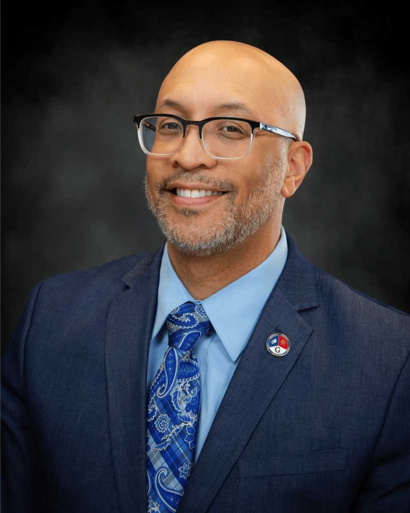 Professional portrait of a smiling man wearing glasses, a blue suit, and a patterned tie, with a badge on his lapel, set against a dark background.