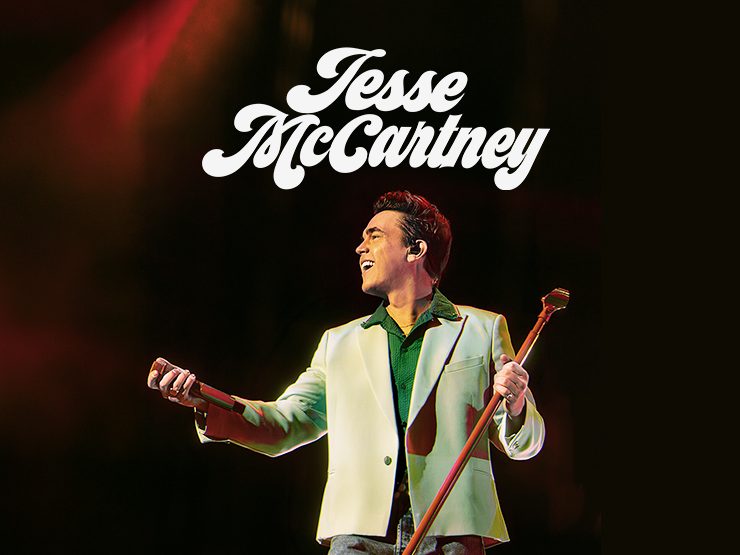 Jesse McCartney performing on stage, smiling and holding a microphone with colorful stage lighting in the background.
